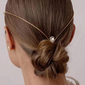 May include: A gold-toned metal headpiece featuring a thin, V-shaped wire band that rests across the back of the head. The center of the band is adorned with a single, clear, pear-shaped crystal pendant that sits just above the nape of the neck.
