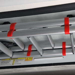 May include: A folded silver aluminum ladder secured with red straps inside a vehicle compartment. The ladder is compact and designed for storage. A black latch and warning label are visible.