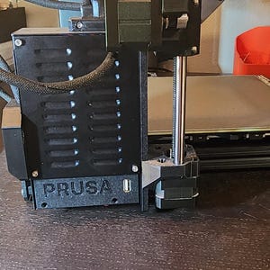 May include: Close-up of a black 3D printer with a silver metal rod and the word "PRUSA" printed on the side.