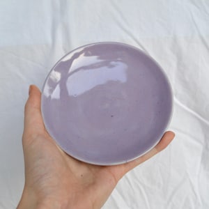 May include: A small, round, lavender-colored ceramic plate. The plate is held in a hand against a white background. The plate has a glossy finish and a simple, minimalist design.