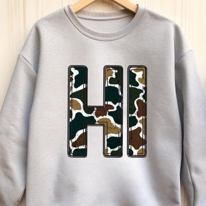 May include: A light gray sweatshirt with the word "HI" in a camouflage pattern. The letters are outlined in black and filled with shades of green, brown, and white. The sweatshirt is hanging on a wooden hanger.