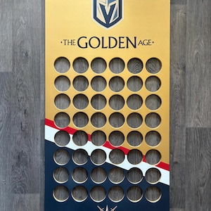 May include: A rectangular gold wooden display board featuring the Vegas Golden Knights logo at the top, the text THE GOLDEN AGE, and a grid of forty-two circular cutouts. The bottom section has diagonal red, white, and navy stripes with a star logo.