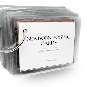 May include: A stack of rectangular cards held together by a silver metal ring. The top card is white with black text that reads NEWBORN POSING CARDS, 50 Poses for Photographers, and FIELDS LANE DESIGN. Each card is enclosed in a clear plastic sleeve.