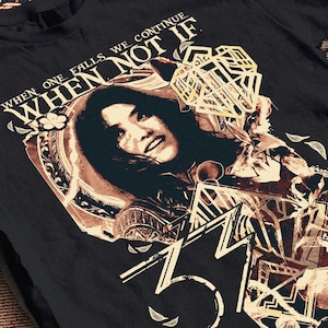 Pode incluir: T-shirt preta com um design gráfico. O design inclui um retrato de mulher e o texto "WHEN ONE FALLS WE CONTINUE WHEN NOT IF". A obra de arte também apresenta formas geométricas e o número "37".
