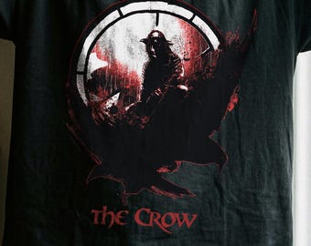 May include: A black short-sleeve t-shirt featuring a graphic print of a figure in a hat and long coat standing within a circular frame, with a large bird silhouette below and the red text "THE CROW" printed underneath the central design.