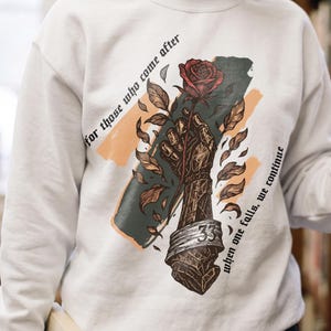 May include: A white crewneck sweatshirt featuring a graphic print of a metallic armored hand holding a red rose. The design includes the text "for those who come after" and "when one falls, we continue" with a number 33 on the wrist cuff.
