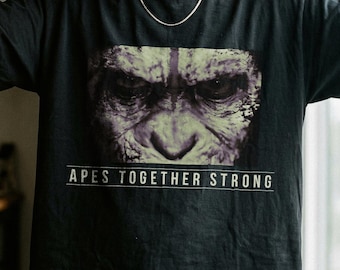 Könnte beinhalten: Ein schwarzes Kurzarm-T-Shirt mit einem Grafikdruck eines Primatengesichts in Grün- und Grautönen. Unter der Grafik ist der Text APES TOGETHER STRONG in weißen Großbuchstaben zwischen zwei horizontalen Linien auf der Brust aufgedruckt.