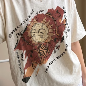 Może przedstawiać: Kremowy t-shirt z krótkim rękawem z nadrukiem graficznym przedstawiającym stylizowaną twarz słońca z geometrycznymi wzorami w odcieniach czerwieni i czerni. Tekst "Losing a rock is better than never having one!" jest nadrukowany czcionką szeryfową.