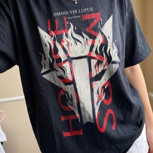Op de afbeelding: Een zwart t-shirt met korte mouwen en een grote grafische print van een brandende fakkel in het midden van een geometrische stervorm. De tekst "OMNIS VIR LUPUS" en "HAIL REAPER" staat boven de afbeelding, met rode letters in het ontwerp.
