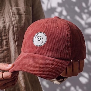 Puede incluir: Una gorra de béisbol de pana color óxido con un parche circular bordado en blanco en la parte delantera. El parche muestra un diseño gráfico negro estilizado que se asemeja a una ola o un remolino. La gorra tiene una corona estructurada.