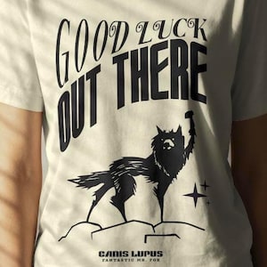 Puede incluir: Camiseta color crema con el texto "GOOD LUCK OUT THERE" en una fuente audaz y estilizada. Debajo del texto hay una ilustración negra de un lobo aullando a una estrella. La camiseta también tiene el texto "CANIS LUPUS FANTASTIC MR. FOX".