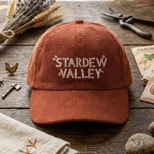 Sorgu başarılı

Gemini şunu dedi:

A rust-colored, corduroy baseball cap sits centered on a rustic wooden table. The front features "STARDEW VALLEY" embroidered in white, blocky letters with small leaf accents. The cap has a classic 6-panel structure and a curved brim.