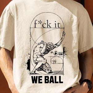 Op de afbeelding: Een beige t-shirt met korte mouwen en een zwarte grafische print van een figuur in een knielende houding binnen een cirkelvormig kader. De tekst "f*ck it" staat boven de figuur en "WE BALL" staat onderaan in dikke zwarte letters.
