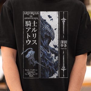 Könnte beinhalten: Ein schwarzes Kurzarm-T-Shirt mit einem Grafikdruck von Artorias the Abysswalker. Das Design enthält japanischen Text, eine Schwertillustration und den englischen Text: Artorias the Abysswalker, Soon, I will be consumed by them, by the Dark.