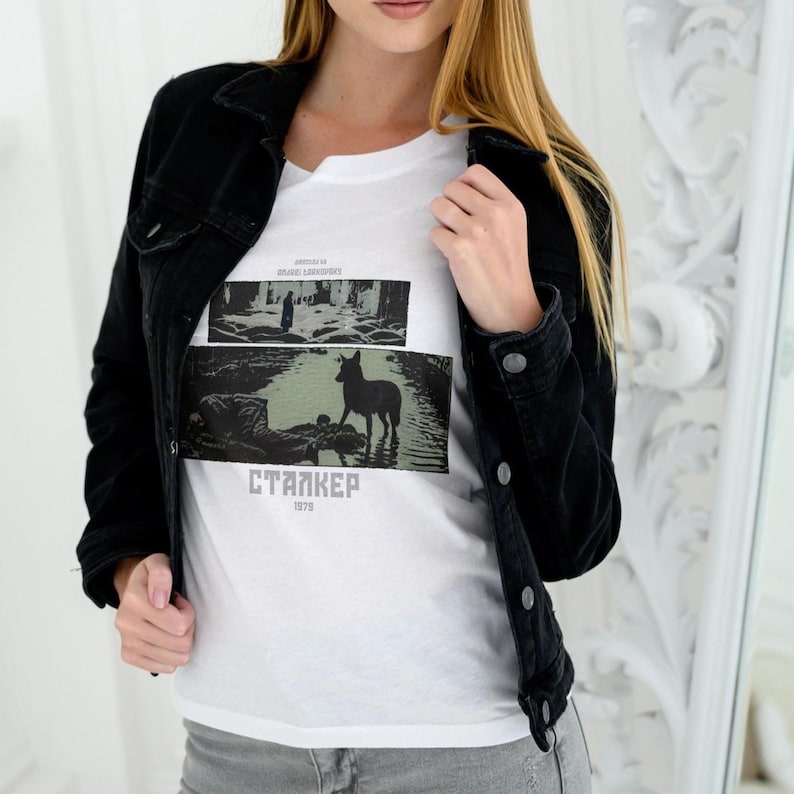 May include: A black denim jacket worn over a white t-shirt featuring a graphic print. The print includes a deer in water, text that reads "STALKER 1979", and other text. The model is wearing gray pants.