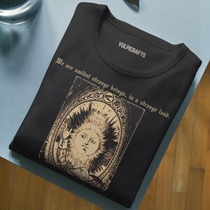 Puede incluir: Una camiseta negra con un estampado gr&aacute;fico en beige de un sol con rostro humano dentro de un marco arqueado. El texto "We are amidst strange beings, in a strange land." aparece impreso encima y "VULPICRAFTS" en el interior del cuello.