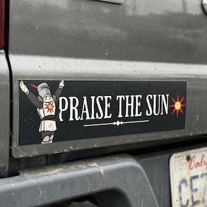Könnte beinhalten: Schwarzer rechteckiger Aufkleber mit dem Text "PRAISE THE SUN" in Weiß. Auf der linken Seite ist ein Cartoon-Ritter mit erhobenen Armen zu sehen. Rechts befindet sich eine rote und gelbe Sonnengrafik. Der Aufkleber ist an einem grauen Fahrzeug angebracht.