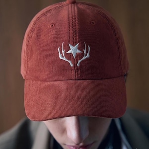 May include: A rust-coloured corduroy baseball cap featuring an embroidered white design on the front panel. The embroidery depicts a five-pointed star positioned directly between two symmetrical, upward-curving deer antlers in a clean white thread.