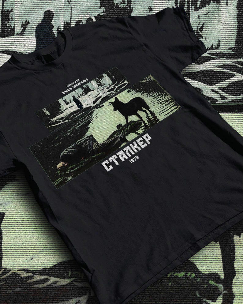 May include: Black t-shirt featuring a graphic from the film "Stalker" with the text "Stalker 1979". The graphic depicts a silhouette of a dog in water and figures in a forest scene. The shirt has a crew neck.