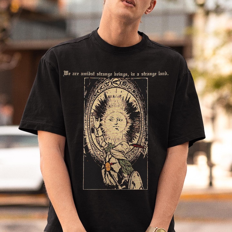 Puede incluir: Una camiseta negra de manga corta con un estampado gr&aacute;fico de un caballero con armadura y un emblema de sol en el pecho, frente a un gran sol con rostro dentro de un marco ovalado. El texto "We are amidst strange beings, in a strange land." est&aacute; impreso encima.