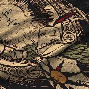 Puede incluir: Una prenda de tela negra con una serigraf&iacute;a en tono dorado que muestra un sol con rostro humano, un caballero con armadura y patrones circulares decorativos. El dise&ntilde;o incluye acentos de follaje verde y un peque&ntilde;o detalle rojo.