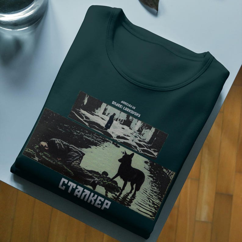 May include: Dark teal t-shirt featuring a graphic from the film "Stalker." The design includes a black silhouette of a dog, a figure in the water, and the film's title in white Cyrillic letters. The text "Directed by Andrei Tarkovsky" is also present.