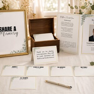 May include: A memorial memory table set featuring a gold-framed sign reading Share a Memory, a wooden box with blank cards, a folded funeral program for Matthew Johnson, a thank you card, five lined prompt cards, and a silver pen on a white cloth.