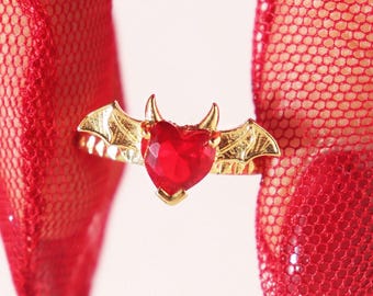 Peut inclure: Une bague en or avec une pierre rouge en forme de cœur et des ailes de chauve-souris. La bague est portée sur un doigt avec un gant en maille rouge.