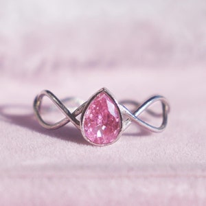 Puede incluir: Un anillo de plata con una piedra preciosa rosa en forma de pera engastada en un delicado diseño de infinito. El anillo se muestra sobre una superficie de terciopelo rosa.
