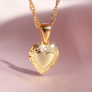 Puede incluir: Un collar de oro con un dije en forma de corazón sobre un fondo de tela rosa.