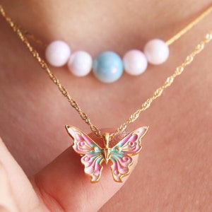 May include: A gold chain necklace with a pink and blue butterfly pendant. The chain also has a row of white and blue beads.