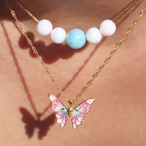 May include: A gold chain necklace with a pink and blue butterfly pendant. A second gold chain necklace with white and blue beads is layered on top.