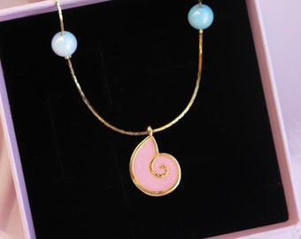 May include: A gold chain necklace with a pink seashell pendant and two light blue beads. The necklace is displayed in a pink box with a black interior.