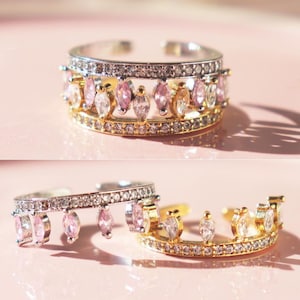 Könnte beinhalten: Zwei offene Ringe mit silbernen und goldenen Details. Ein Ring hat rosa und klare Edelsteine, der andere hat hängende rosa Edelsteine. Die Ringe sind vor einem hellrosa Hintergrund platziert.