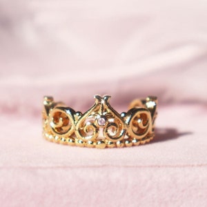 Pode incluir: Um anel de coroa dourado com uma única pedra transparente no centro, descansando sobre uma caixa de veludo rosa.