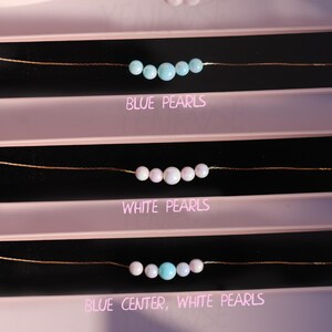 May include: Three necklaces with different pearl combinations. The top necklace has blue pearls, the middle necklace has white pearls, and the bottom necklace has white pearls with a blue pearl in the center. The necklaces are displayed on a pink background with the text "Blue Pearls", "White Pearls", and "Blue Center, White Pearls" underneath each necklace.