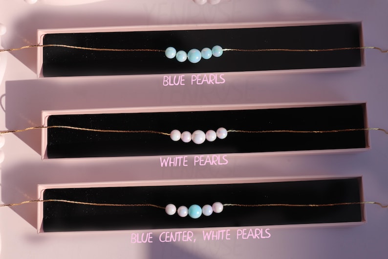 May include: Three necklaces with different pearl combinations. The top necklace has blue pearls, the middle necklace has white pearls, and the bottom necklace has white pearls with a blue pearl in the center. The necklaces are displayed on a pink background with the text "Blue Pearls", "White Pearls", and "Blue Center, White Pearls" underneath each necklace.