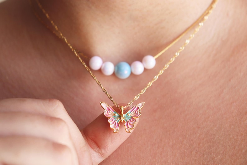 May include: A gold chain necklace with a pink and blue butterfly pendant. The chain also has a row of white and blue beads.