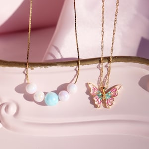 May include: Two gold necklaces on a pink surface. One necklace has a butterfly pendant with pink, green, and white enamel. The other necklace has a row of pastel-colored beads.