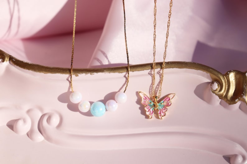 May include: Two gold necklaces on a pink surface. One necklace has a butterfly pendant with pink, green, and white enamel. The other necklace has a row of pastel-colored beads.