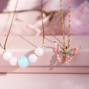 May include: Two gold necklaces with pendants. One necklace has a pastel blue and white pearl design. The other necklace has a pink and green butterfly pendant.