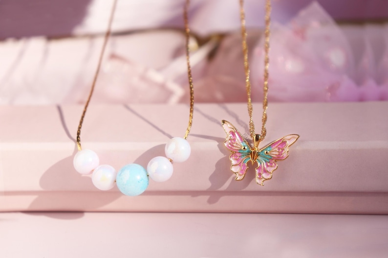 May include: Two gold necklaces with pendants. One necklace has a pastel blue and white pearl design. The other necklace has a pink and green butterfly pendant.