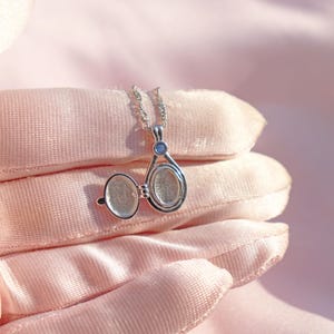 May include: A silver locket necklace with an oval pendant, open to reveal a space for a photo. The locket hangs from a delicate chain and features a small blue gemstone. The locket is displayed on a pink, textured surface.