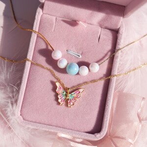 May include: Two gold necklaces with a butterfly pendant and pearl and blue beads in a pink velvet jewelry box.