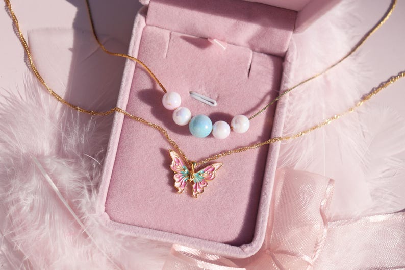 May include: Two gold necklaces with a butterfly pendant and pearl and blue beads in a pink velvet jewelry box.