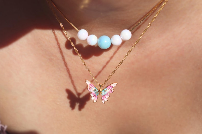 May include: A gold chain necklace with a pink and blue butterfly pendant. A second gold chain necklace with white and blue beads is layered on top.