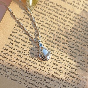 May include: A silver necklace with a teardrop-shaped pendant, featuring a pearlescent center stone and small blue accents. The necklace rests on an open book with printed text, creating a vintage aesthetic.
