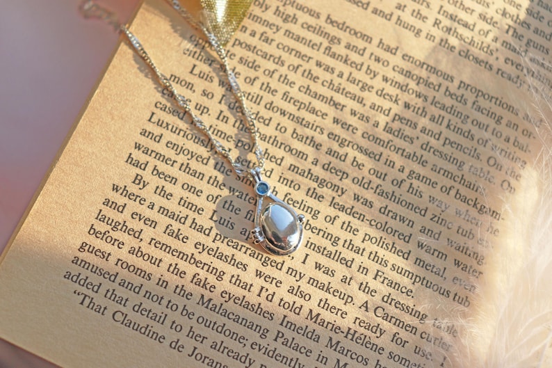 May include: A silver necklace with a teardrop-shaped pendant, featuring a pearlescent center stone and small blue accents. The necklace rests on an open book with printed text, creating a vintage aesthetic.