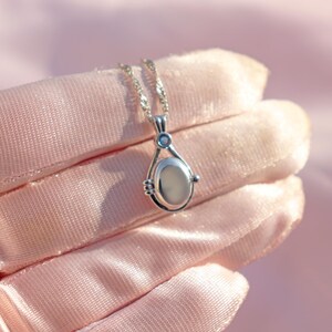 May include: A silver pendant necklace with a teardrop-shaped locket. The locket has a light-colored oval center and a small blue accent stone. The necklace is displayed on a pink satin surface.
