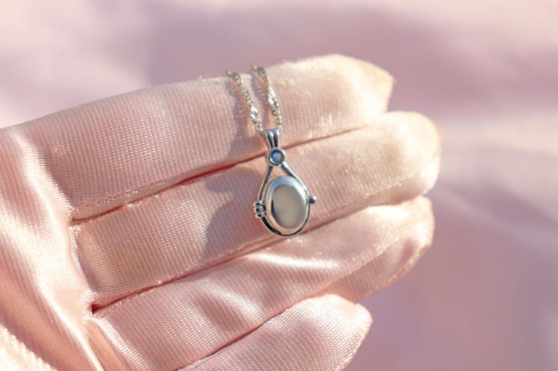 May include: A silver pendant necklace with a teardrop-shaped locket. The locket has a light-colored oval center and a small blue accent stone. The necklace is displayed on a pink satin surface.